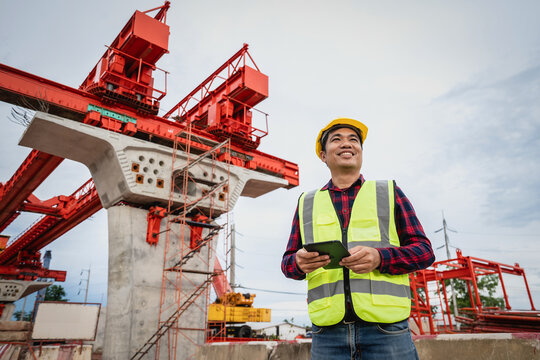Asian civil engineers use tablets to record electronic data at a concrete highway bridge construction site. A male supervisor is inspecting things at the new concrete road and bridge construction site