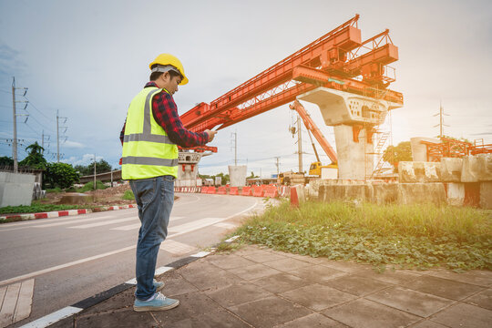 Construction foreman and engineer, wearing safety equipment, are looking at blueprints at a high bridge construction site where large cranes are operating.