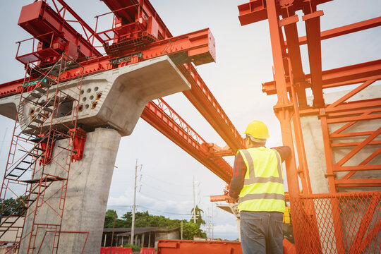 Construction foreman and engineer, wearing safety equipment, are looking at blueprints at a high bridge construction site where large cranes are operating.