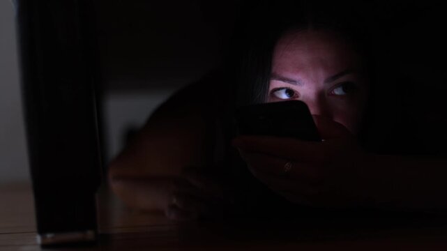 Female aggressor hides beneath bed as police arrive. She uses smartphone while flashing emergency lights expose her frightened face.