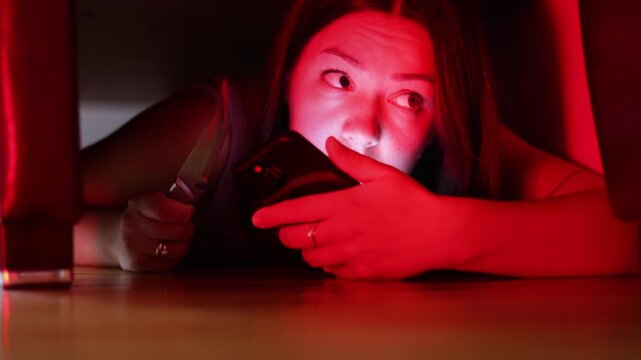 Woman hides beneath bed while speaking with police by phone. Kitchen knife remains in her hand as flashing lights illuminate her frightened face.