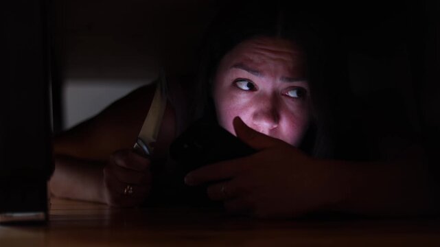 Female criminal hides beneath bed while police search nearby. She grips kitchen knife and uses smartphone to contact accomplices.