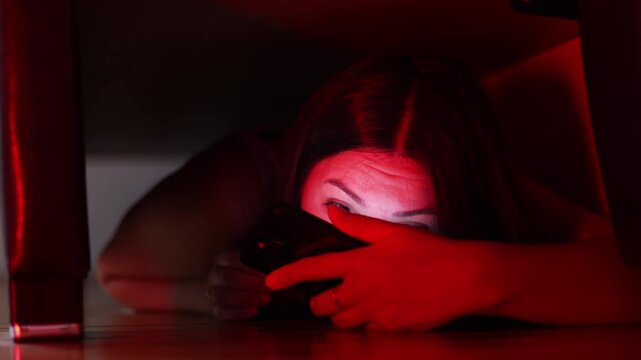 Frightened woman hides beneath bed and cautiously looks outside. Smartphone connects her with police while flashing lights illuminate her anxious face.