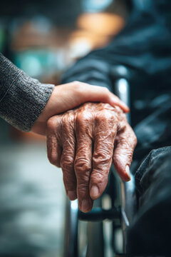 An elderly man sits in a wheelchair while a young woman gently holds his hand. The background is blurred, showing a caring atmosphere in an indoor setting