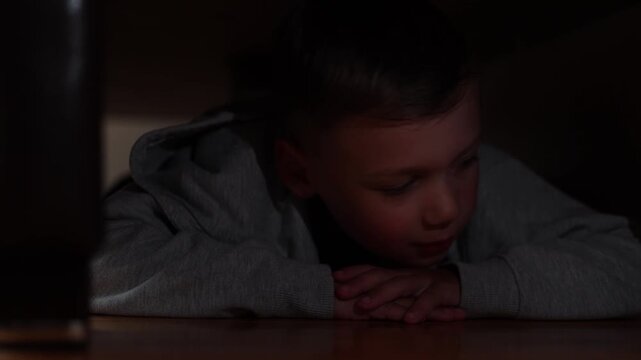 Six year old boy hides beneath bed after making prank police call. Flashing police lights illuminate his face while he waits in confusion.