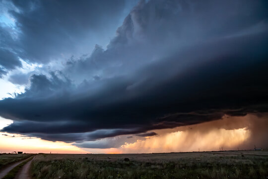 View of a massive supercell storm cloud with heavy rain curtains and dramatic sunset light over a vast open field.