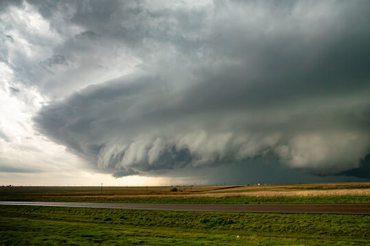 View of a massive supercell storm cloud looming over a flat rural landscape with a road in the foreground.