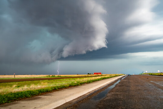 View of a supercell storm with a lightning bolt striking over a rural landscape with green fields and a distant red building under dark, ominous clouds.