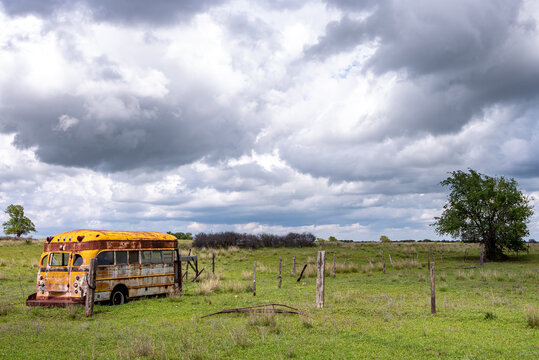 View of an abandoned, rusted yellow school bus sitting in a grassy field under a dramatic, cloudy sky with wooden fence posts and a lone tree in the background.