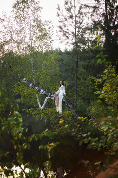 woman relaxes on a thick tree branch over a lake. sun shines through the leaves as she enjoys the quiet nature around her. Tall trees rise in the background