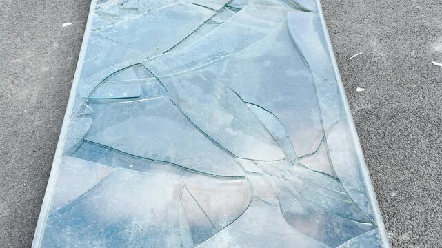 A large sheet of glass shatters into sharp cracks on a dark asphalt street. This broken pane creates a moody, tense feeling of destruction and unexpected danger.