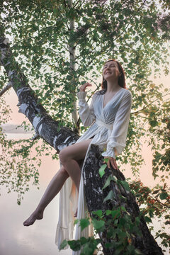 woman sits on a tree branch stretching over a lake. She smiles and enjoys the fresh air and sunlight as the sky shows soft colors of sunrise in the background