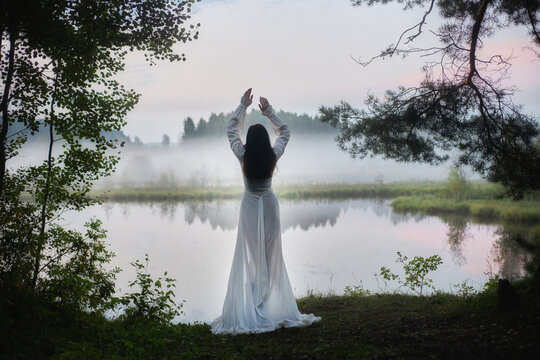 woman stands by a river with mist rising around her. She is wearing a long white dress and raises her arms. sun is just rising, and the scene is filled with trees and water