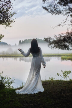 woman in a white dress stands by a body of water at dawn. Fog hovers over the water and trees are visible in the background. scene shows a quiet moment in nature