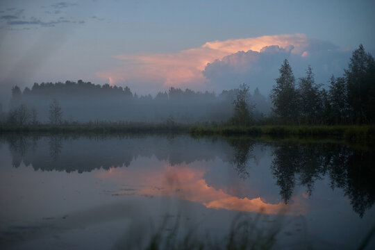 Morning light brightens the sky with colors over a lake. Trees line the shore and mist lingers over the water. scene shows a peaceful start to the day