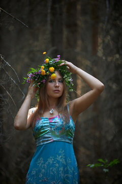 woman is standing in a forest wearing a dress and a flower crown. She is holding the crown while looking ahead. Sunlight filters through the trees