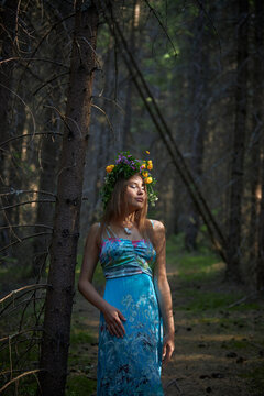woman is in a forest wearing a blue dress and a crown made of flowers. She appears to be standing still among trees. Sunlight comes through the branches