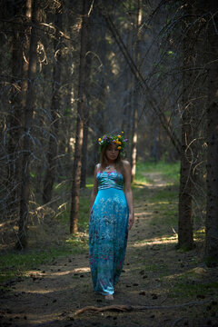 woman walks on a dirt path surrounded by tall trees. She wears a long dress and a flower crown. Sunlight filters through the leaves of the forest while she moves gracefully