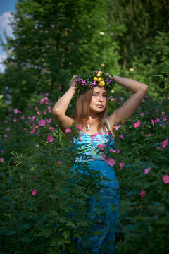 woman stands in a garden filled with colorful flowers. She wears a crown made of flowers and poses with her hands raised. sun shines brightly in the afternoon sky
