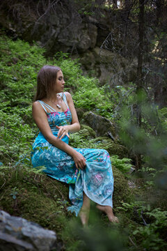 woman rests on a rock in a forest area. She wears a long, colorful dress and looks thoughtful. greenery surrounds her, offering a natural background. Sunlight filters through trees