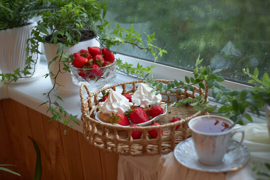 basket holds strawberries topped with whipped cream placed on a table by a window. cup rests nearby, and greenery is visible outside the glass. Rain falls against the window