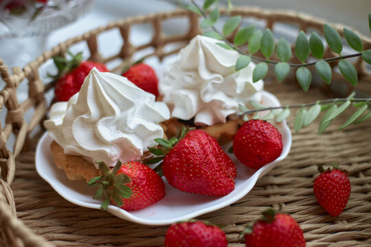 Two desserts topped with whipped cream are placed on a plate with red strawberries. setting is casual and warm, with a woven basket underneath