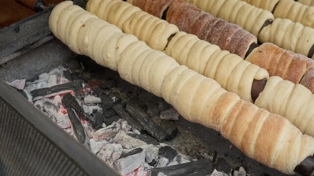 Traditional chimney cake pastry rotates on a spit over burning charcoal coals on a street grill. Sweet dough bakes into a delicious sugar dessert at an authentic European food festival.