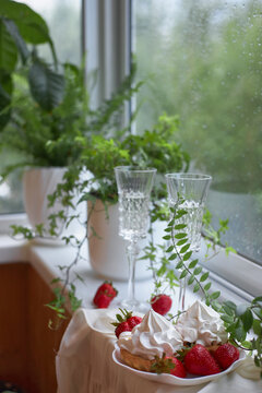 Two clear glasses filled with sparkling drink stand next to a bowl of dessert topped with whipped cream and strawberries. setting features indoor plants by the window