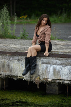 young woman in casual clothing sits on the edge of an old dock by a body of water. She looks contemplative while the sun shines in a summer environment