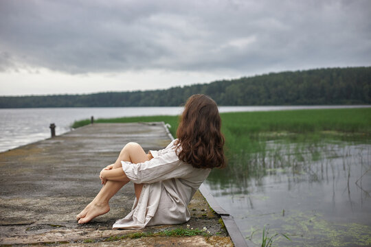 woman is sitting at the edge of a dock by a lake. She is gazing out at the water and the green plants around. sky is cloudy. It is day time