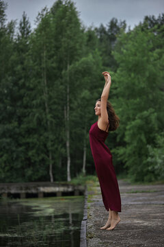 woman stands on the edge of a body of water with her arms raised. She is barefoot and wearing a red dress. Green trees surround her in a cloudy setting