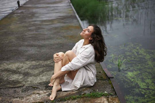 woman is seated on a stone dock with her knees pulled to her chest, gazing up at the sky. calm water reflects greenery around her