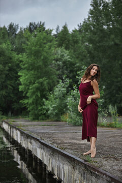 woman poses on a dock at a calm body of water. setting includes greenery and overcast skies. She stands barefoot and has a relaxed stance