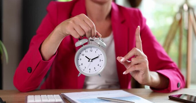 A person in a bright pink jacket holds an old clock while sitting at a desk. The room is lively with plants in the background, symbolizing time management and focus.