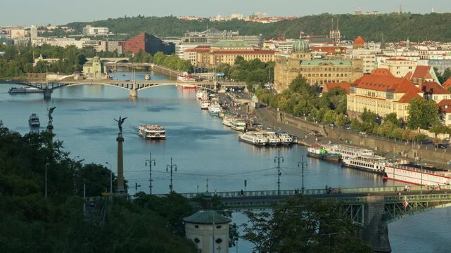 Cruise boats sail along Vltava River past Cech Bridge in historic Prague city. European urban landscape with stone embankment offers scenic background for Czech Republic tourism travel.