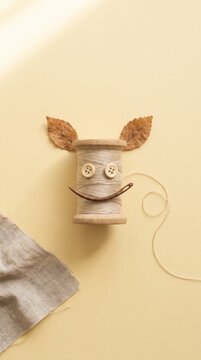 Whimsical thread spool face with oak leaf ears and button eyes on warm cream background