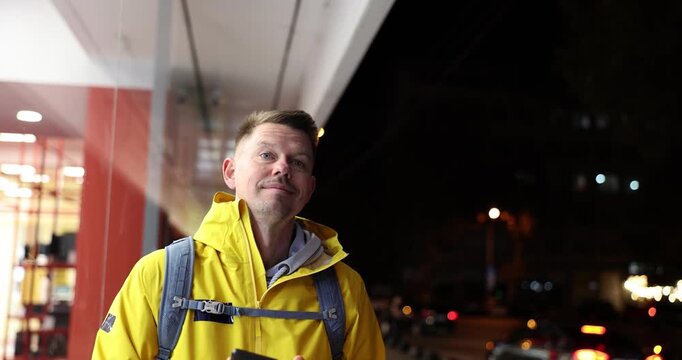 A cheerful man in a bright yellow jacket greets the camera with a warm smile and playful gestures. The lively city lights twinkle behind him, creating a vibrant atmosphere.