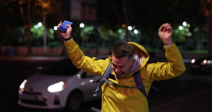 A man wearing a bright yellow jacket joyfully dances in the streets, soaking in the vibrant energy of the nighttime city. His laughter echoes as he embraces the moment with excitement.