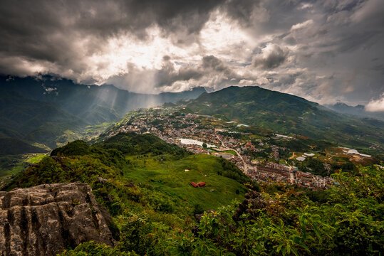 View of Sa Pa town nestled in the lush green mountains of the Hoang Lien Son range under dramatic storm clouds with sunbeams breaking through, Sa Pa, Lao Cai, Vietnam.