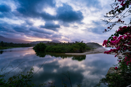 View of Parque Nacional Natural Tayrona featuring a river estuary meeting the sea under a dramatic cloudy sky with vibrant pink bougainvillea in the foreground, Magdalena, Colombia.
