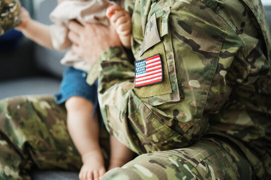 Male american soldier in military uniform hugs his little son at home while baby boy playing with his hat - Emotional happy homecoming concept - Main focus on flag