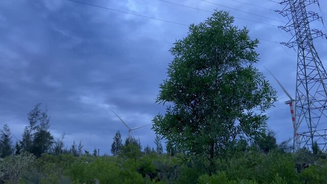Wind turbines spinning at twilight under dramatic stormy sky in Quy Nhon Vietnam with power lines, medium panning shot