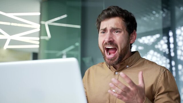 Close-up of angry mature businessman speaking emotionally during an online video call in a modern office. Frustrated professional reacting to a workplace conflict while facing the laptop camera.
