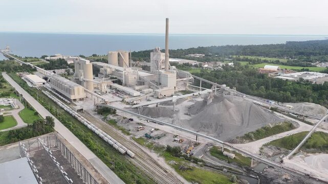 High-angle aerial shot circling the large concrete silos, kiln exhaust stack, raw material aggregate piles, and freight train tracks of a cement factory on Lake Ontario in Mississauga, Canada