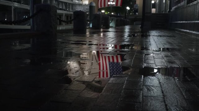 A wet cobblestone walkway at night reflects an American flag hanging above, creating a patriotic urban scene after rainfall.
