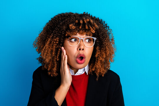 Surprised professional woman with curly hair and glasses reacting in shock against bright blue background