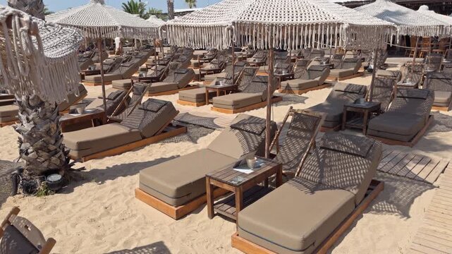 Elevated view captures rows of cushioned sunbeds with wooden side tables under white canopy tents on a sandy beach