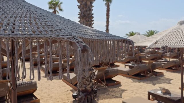 A large woven canopy shelters sun loungers on a sandy beach resort with palm trees and a thatched hut nearby