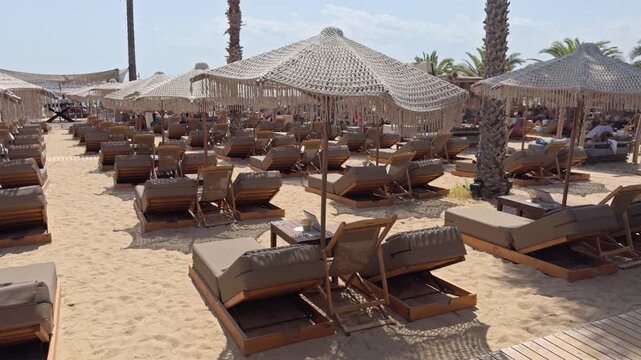 A sandy path leads between symmetrical rows of sun loungers under pointed canopy tents at a beach resort