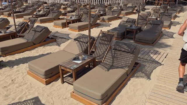 A person walks along a sandy beach past rows of cushioned loungers under canopy tents in a resort